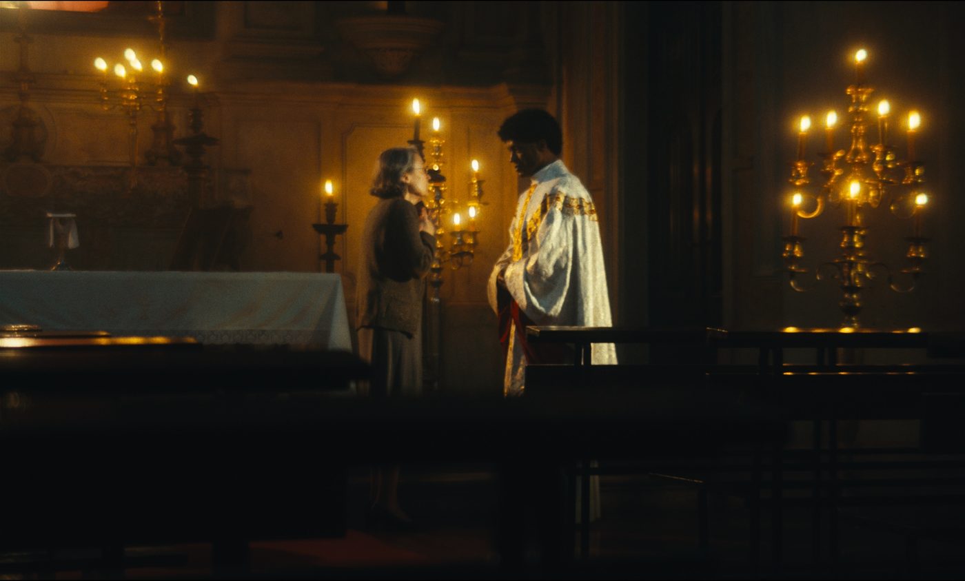 O padre e uma mulher idosa junto ao altar, iluminados por velas